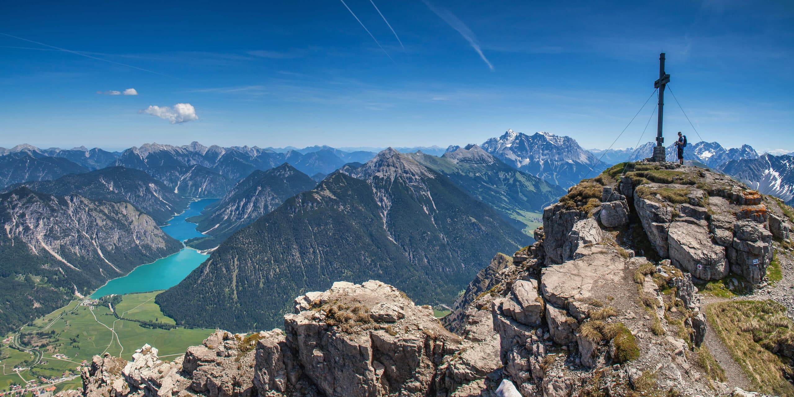 Wanderer am Gipfelkreuz vor leuchender Berglandschaft mit Fluss