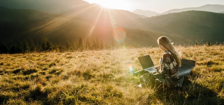 Person sitzt mit einem Laptop auf einer Wiese und arbeitet in der Sonne, umgeben von Bergen und Wäldern.